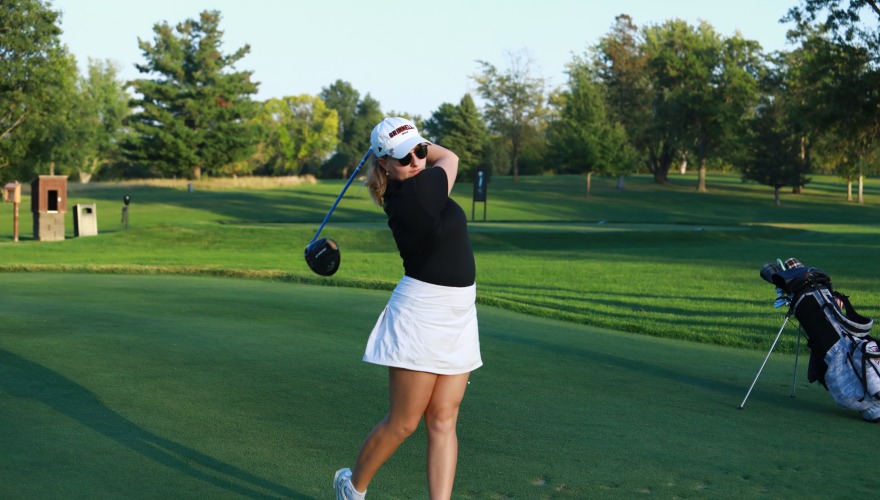 A golfer wearing a black top, white skirt, and white hat follows through after a shot