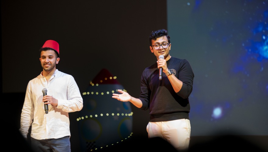 Parikshit on stage with another student wearing a red fez hat