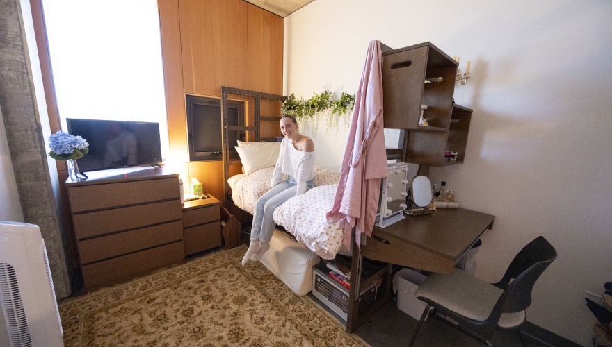 A young woman sits on a raised bed covered in a white bedspread