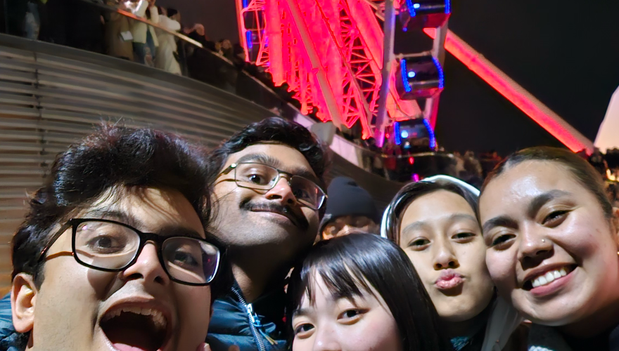 A group of students poses for a selfie in front of a brightly lit ferris wheel at night