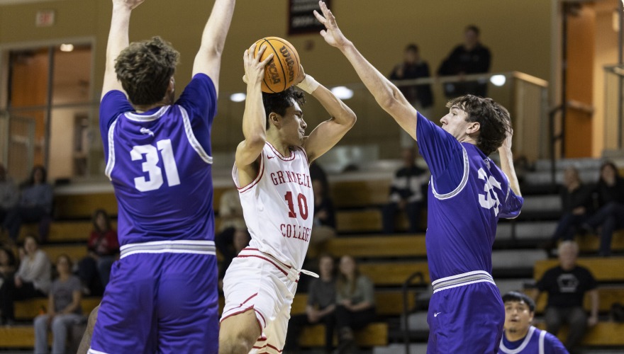A basketball player wearing a white Grinnell uniform jumps with the ball in the midst of two defending players in purple