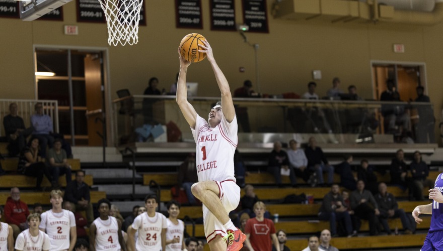 A basketball player in a white Grinnell uniform holds the ball as he jumps to dunk it in the basket