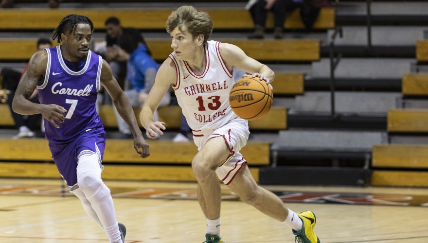 A basketball player with wearing a white Grinnell uniform, number 13, dribbles the ball in front of a defending player in purple