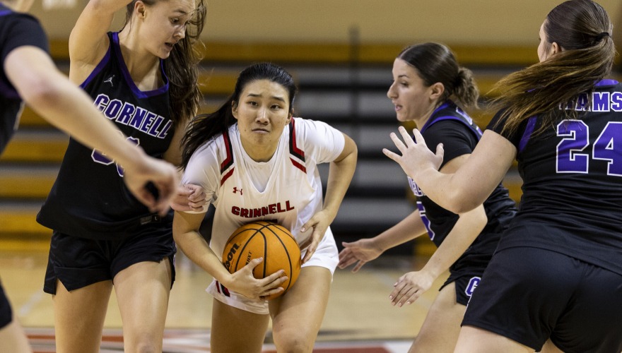 A basketball player with her hair in a dark ponytail and wearing a white Grinnell uniform holds the ball in the midst of three defending players in black