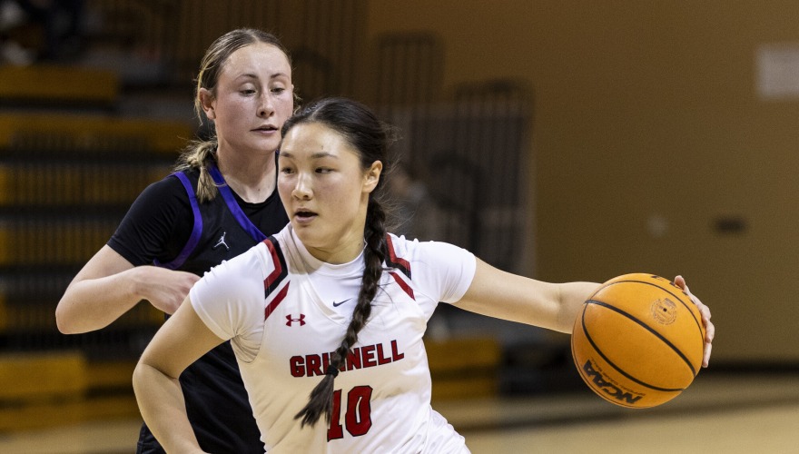 A basketball player with her hair in a dark braid and wearing a white Grinnell uniform, number 14, dribbles the ball in front of a defending player in black