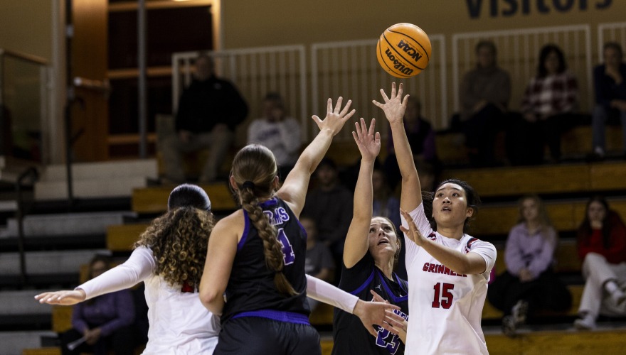A basketball player wearing a white Grinnell uniform reaches for the ball along with two defending players in black