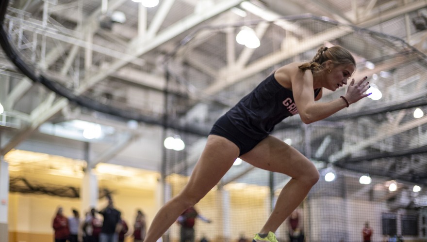 A student with blonde hair and wearing a black track uniform runs on an indoor track