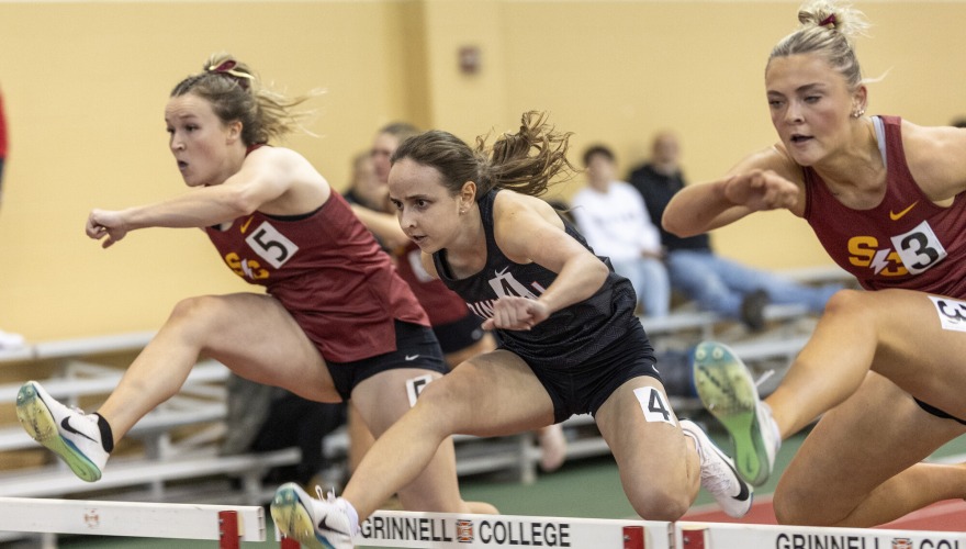 Three students in red and black track uniforms leaping over hurdles