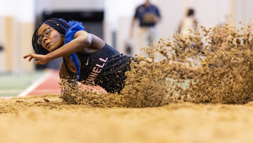 A student with black and blue braids wearing a black Grinnell track uniform lands in the sand pit for long jump