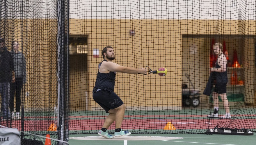 A student wearing a black Grinnell track uniform with his arms outstretched in the middle of a hammer throw