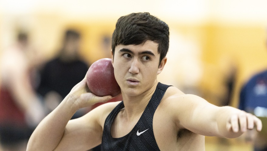 A student with short dark hair and wearing a black Grinnell track uniform holds a shot put behind his head