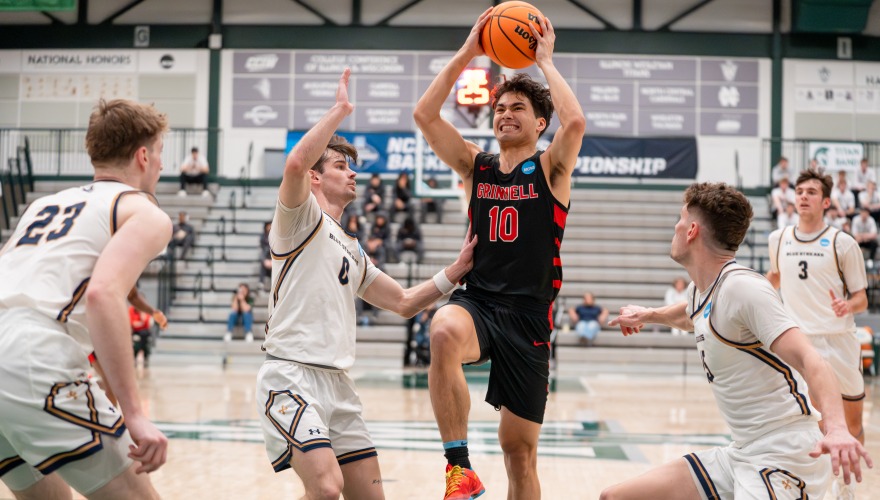 Grinnell basketball player in black holds ball above his head in the midst of three defenders in white