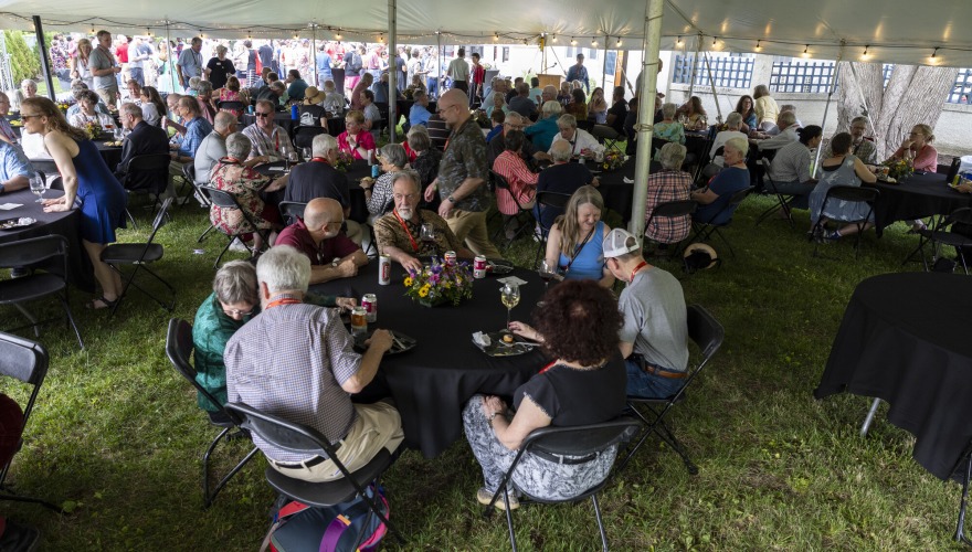 A large group of alumni under a white tent standing and sitting at tables during the President’s Reception.