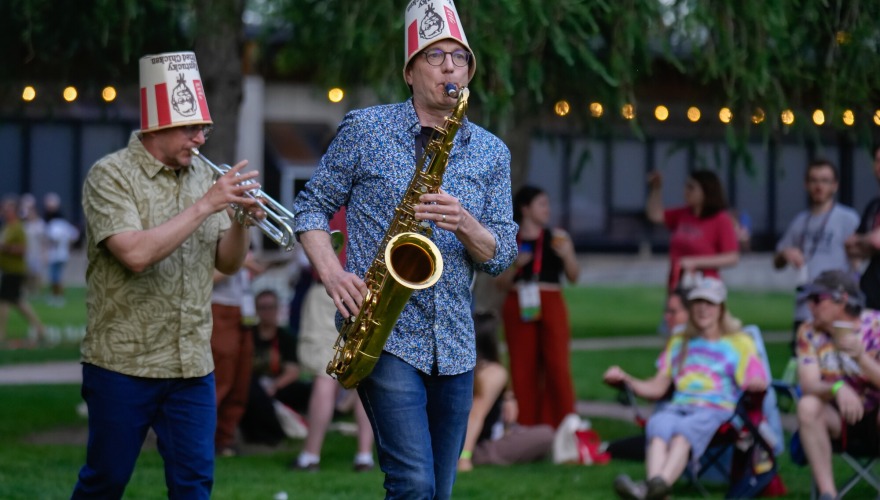 Two alums play the saxophone and trumpet while wearing KFC buckets on their heads and walking through Block Party.