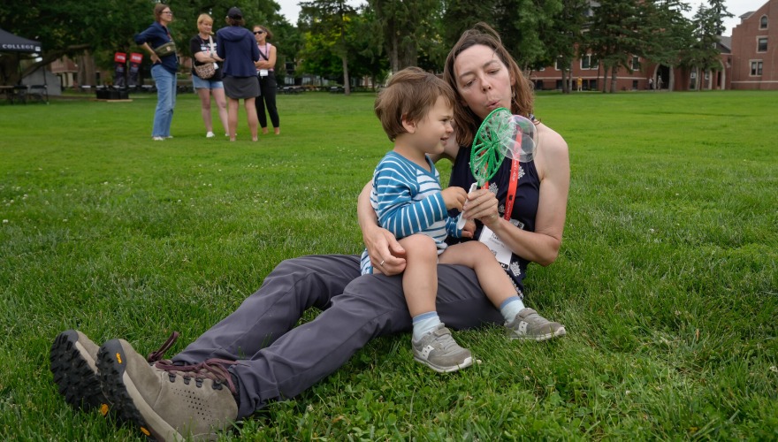 An alum with their child on their lap blow bubbles from a wand on Mac Field.