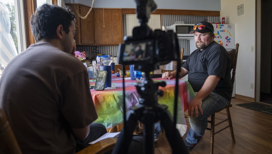 Des Moines Register reporter Nick El Hajj interviews a man at a small dining room table inside the man's house. A digital camera on a tripod is pictured recording the interview.