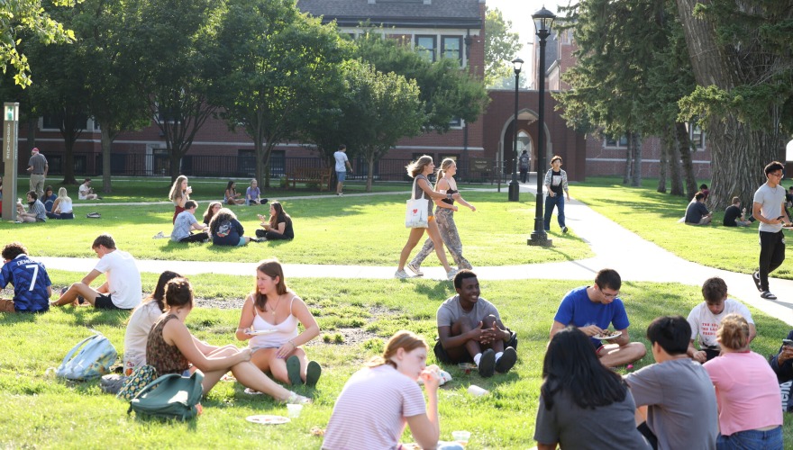 A large group of students sit on the grass eating dinner.