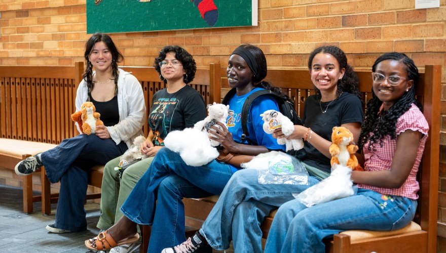 Students in pre-orientation programs sit on a bench and smile as they hold up stuffed animals they've made.