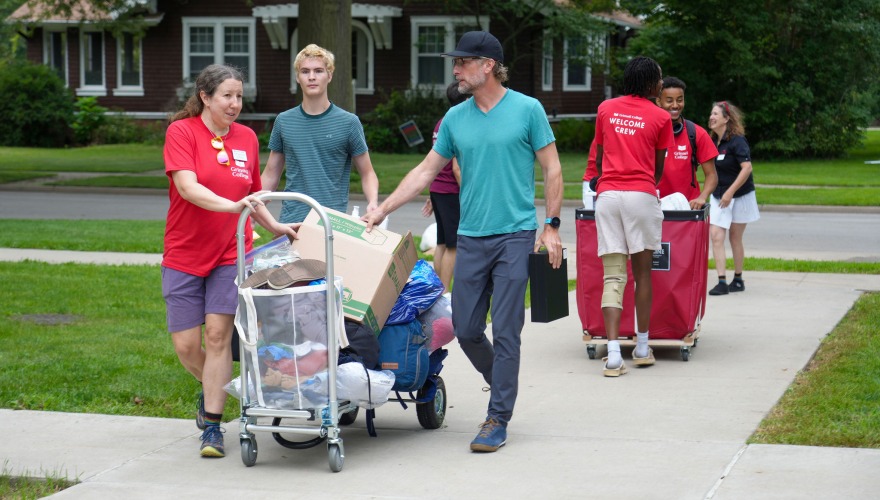 Move-in crew members push carts of items up a sidewalk and chat with an incoming student and their family member.