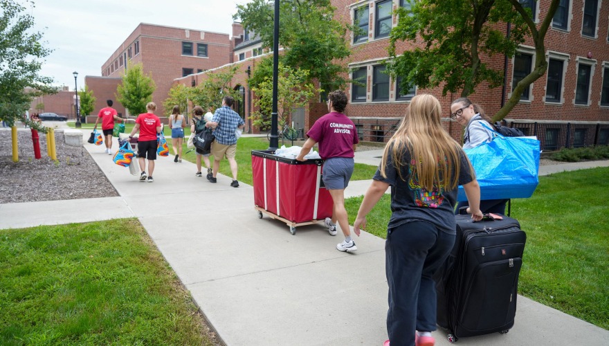 Move-in crew members push several carts on a sidewalk followed by students wheeling luggage.