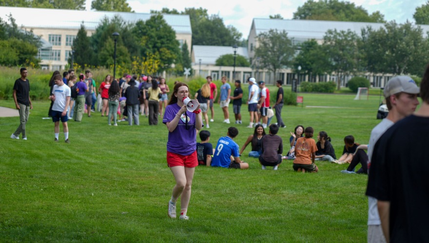 A student mentor talks into a megaphone while small groups of students stand and sit on the grass in the background.