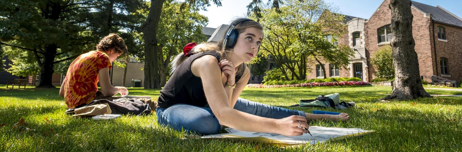 Two students sit in grass, one with headphones looking up