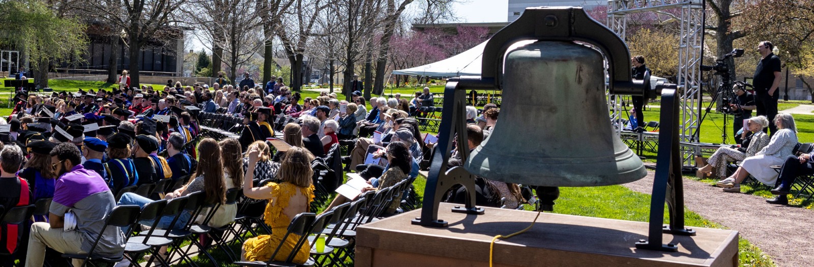 Iowa College Bell in the foreground, with an audience at the commencement stage behind it