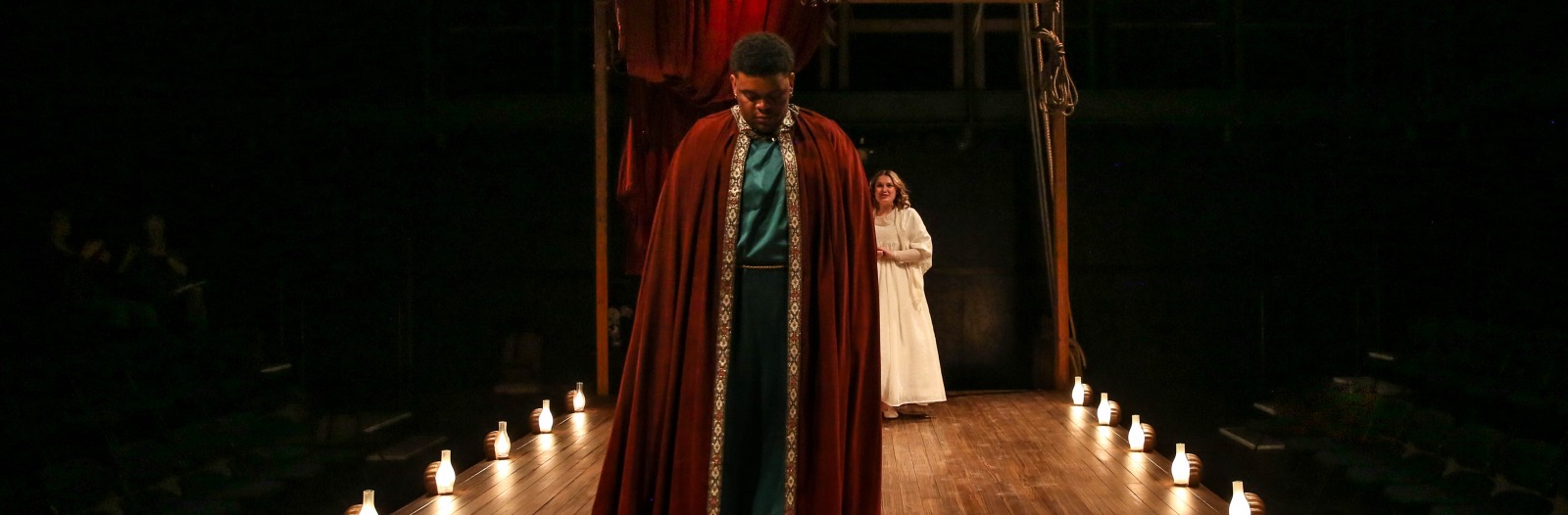 A performer in a red cloak treads between period-style footlights