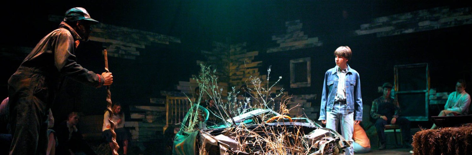Performers in faded rural costuming converse across a beat up car in Flanagan Theatre