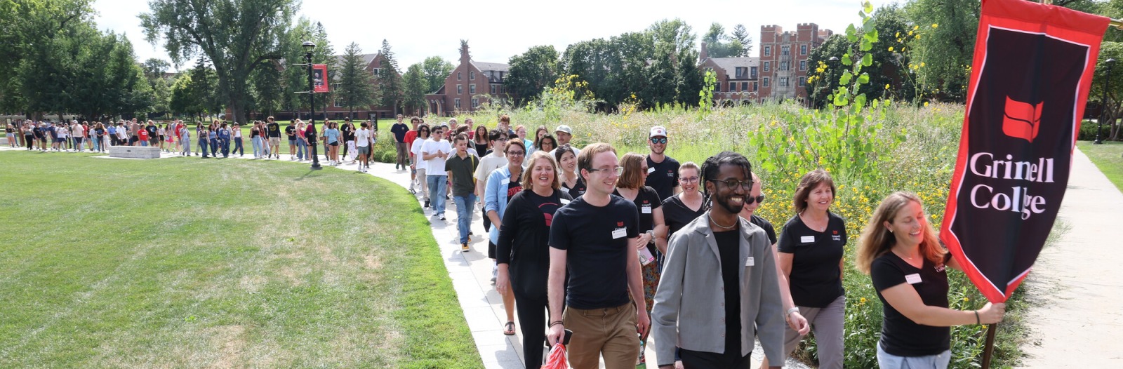 First-year students parade down a sidewalk with one at the beginning of the line holding a sign that reads "Grinnell College"