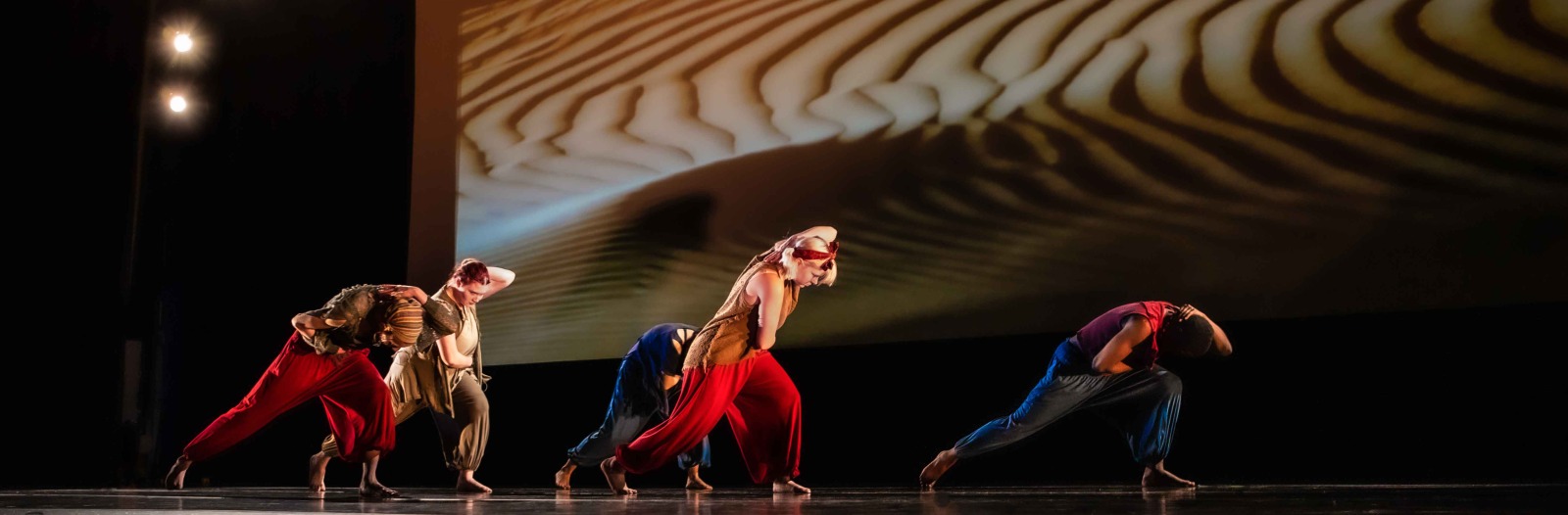 A collections of student performers dance in unison onstage in front of a large projected image of a sand dune. The students wear flowing apparel in jewel tones, and the dance is dramatically lit with a modern feel.