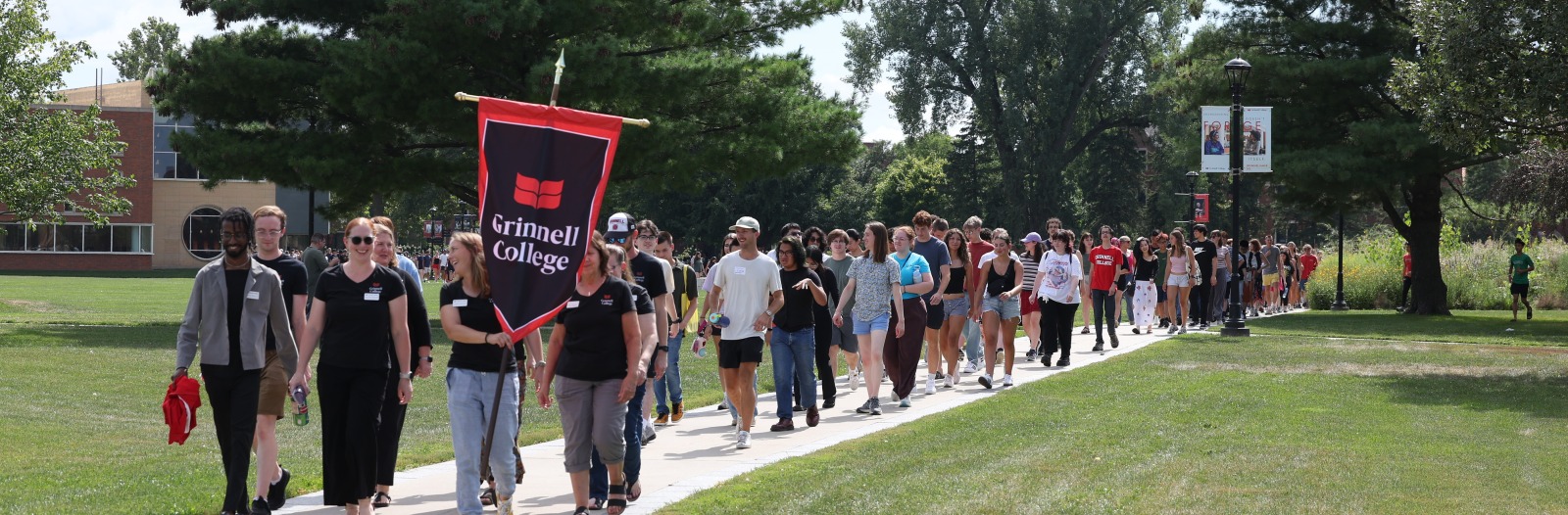 New students are led along a path by an orientation leaders holding a Grinnell College banner.