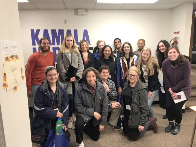 Group of people pose in front of Kamala Harris sign, with a folder taped to the wall marked 'before you leave, register to vote'