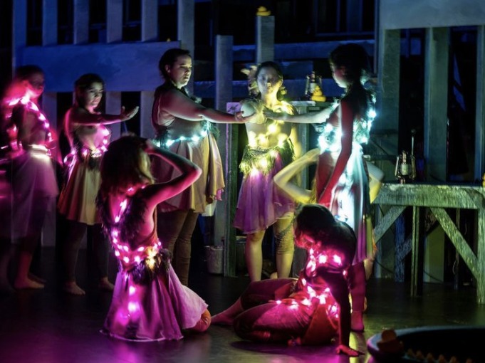 Performers dance with neon lights on their clothes in a dark room.