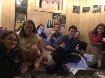 snapshot of seven people - most sitting on a shared bed - smiling and giving thumbsup