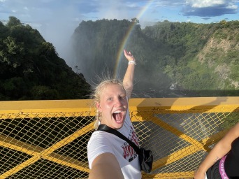 A young woman smiles while standing on a bridge over a deep gorge with a rainbow overhead