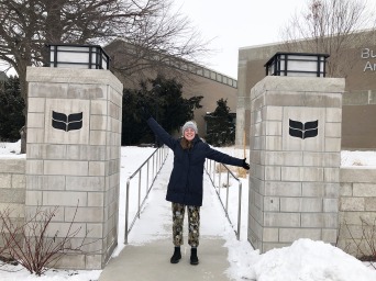 Meredith in between the Grinnell gateposts, smiling and with open arms, with snow on the ground