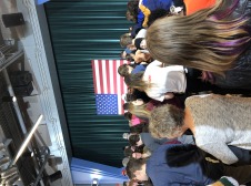 a crowd of adults of all ages standing in front of a stage with the American flag prominently displayed