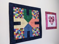 quilted wall hanging depicts four houses in bright colors on a colorful checked field with a black border and a smaller one with heart and border in shades of red on a white field