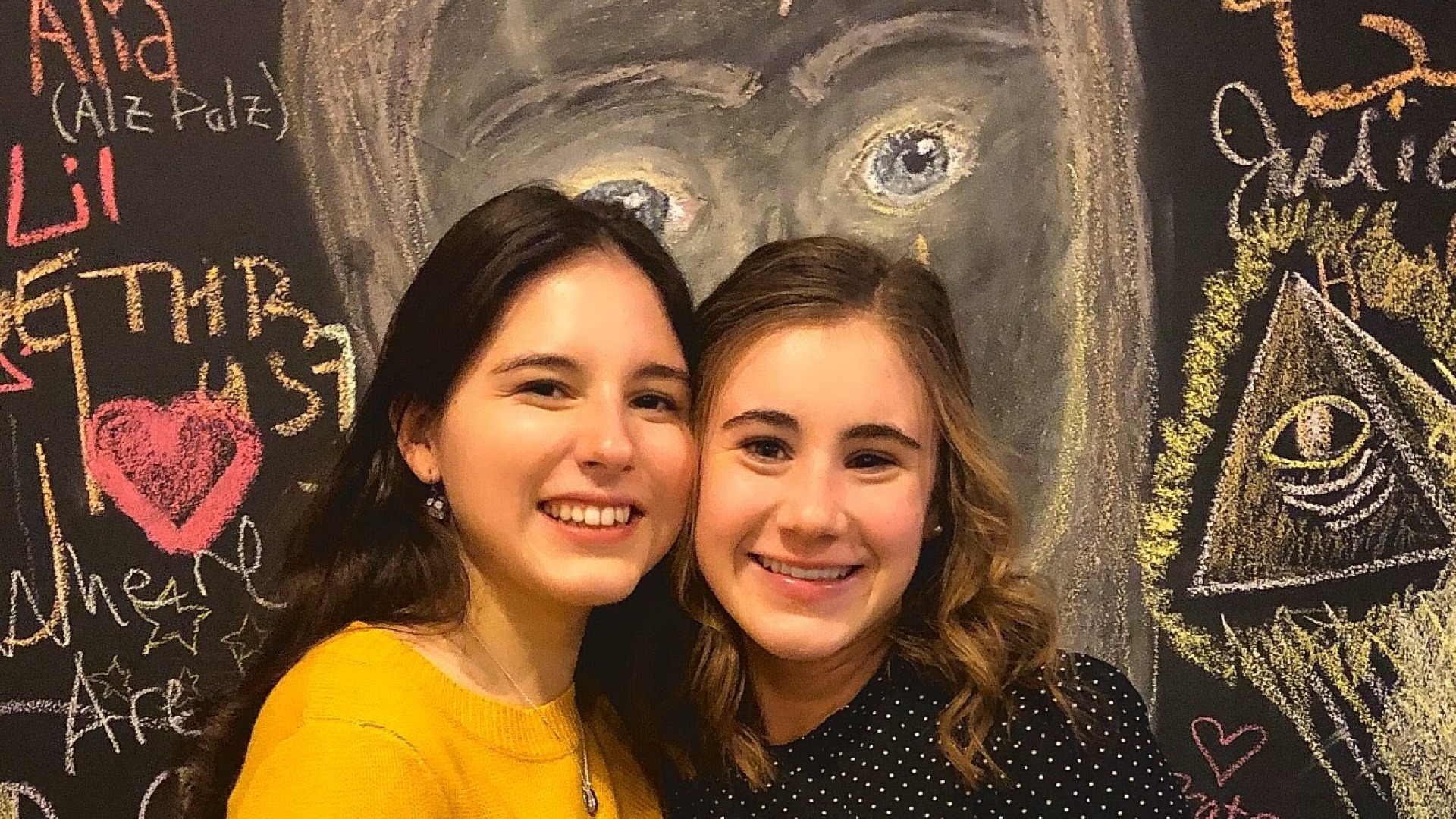 Two girls in photo in front of chalk board.