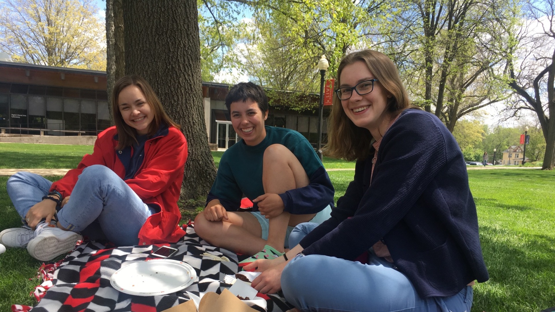Three students share a chocolate bundt cake while sitting on a blanket in the grass