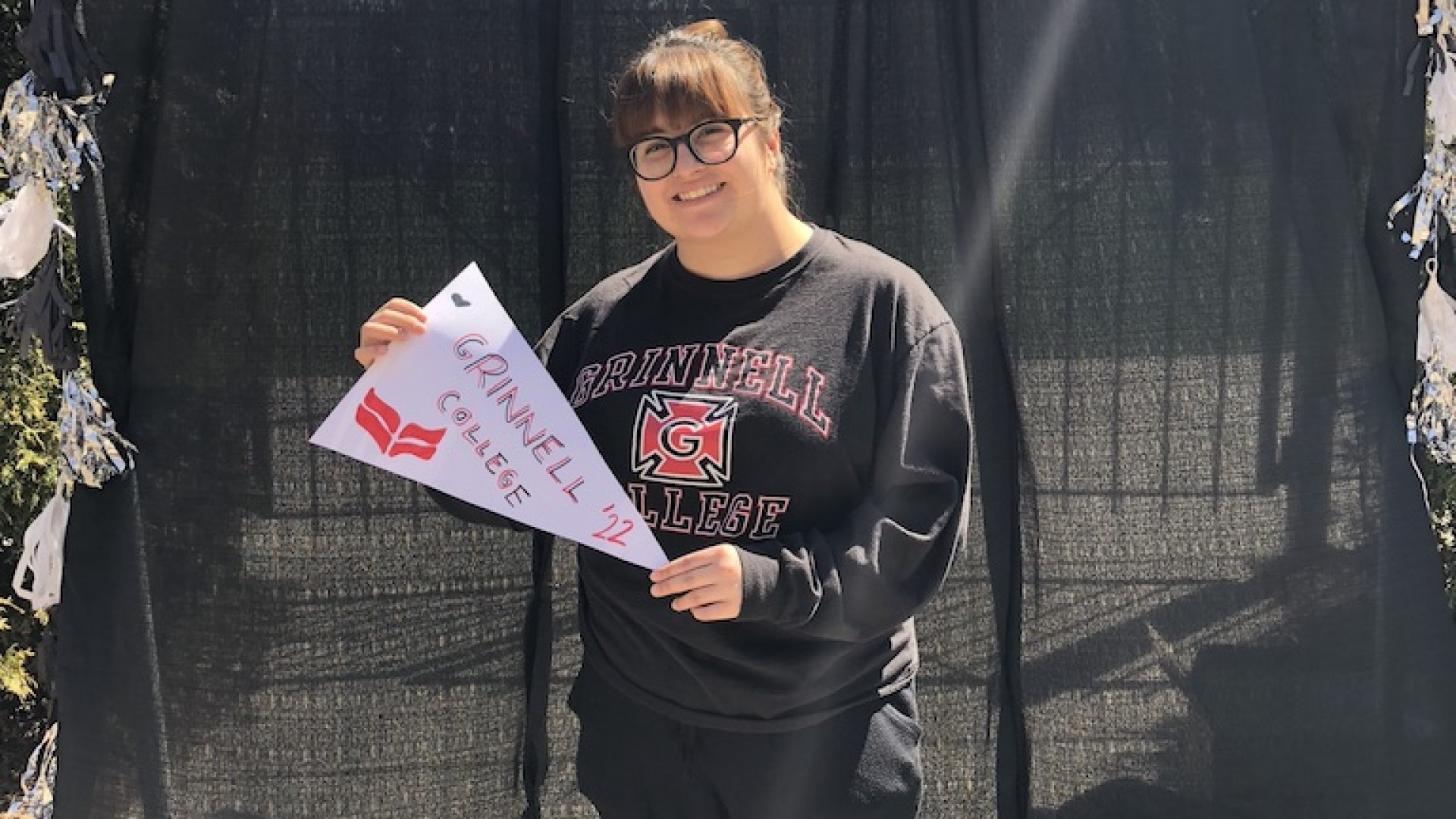Eva in a Grinnell College sweatshirt holding a hand-lettered Grinnell College pennant in front of a background reading Congrats