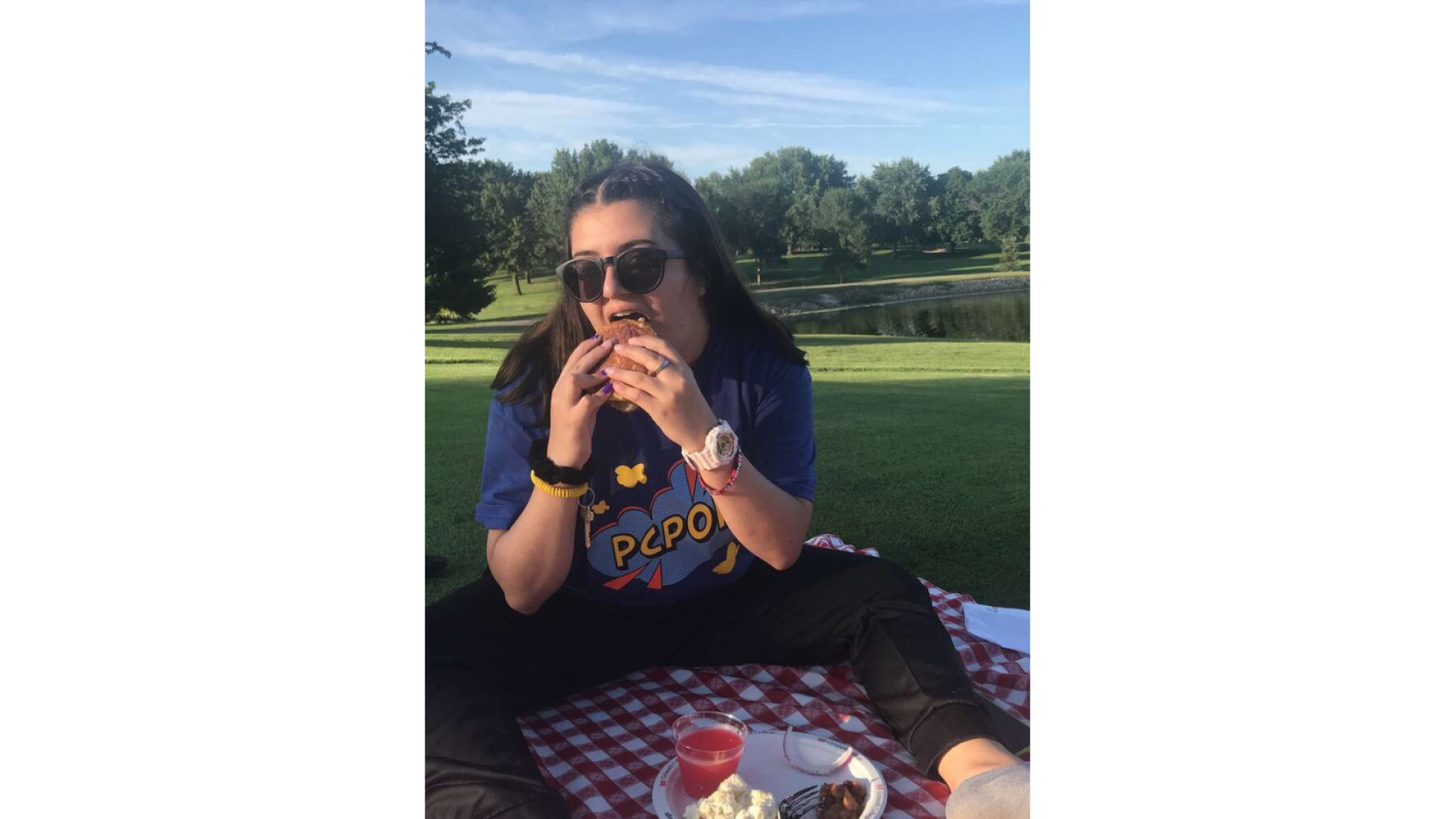 Eva biting into a sandwich at a picnic she is sitting on a red and white checked cloth with the rest of her meal between her legs