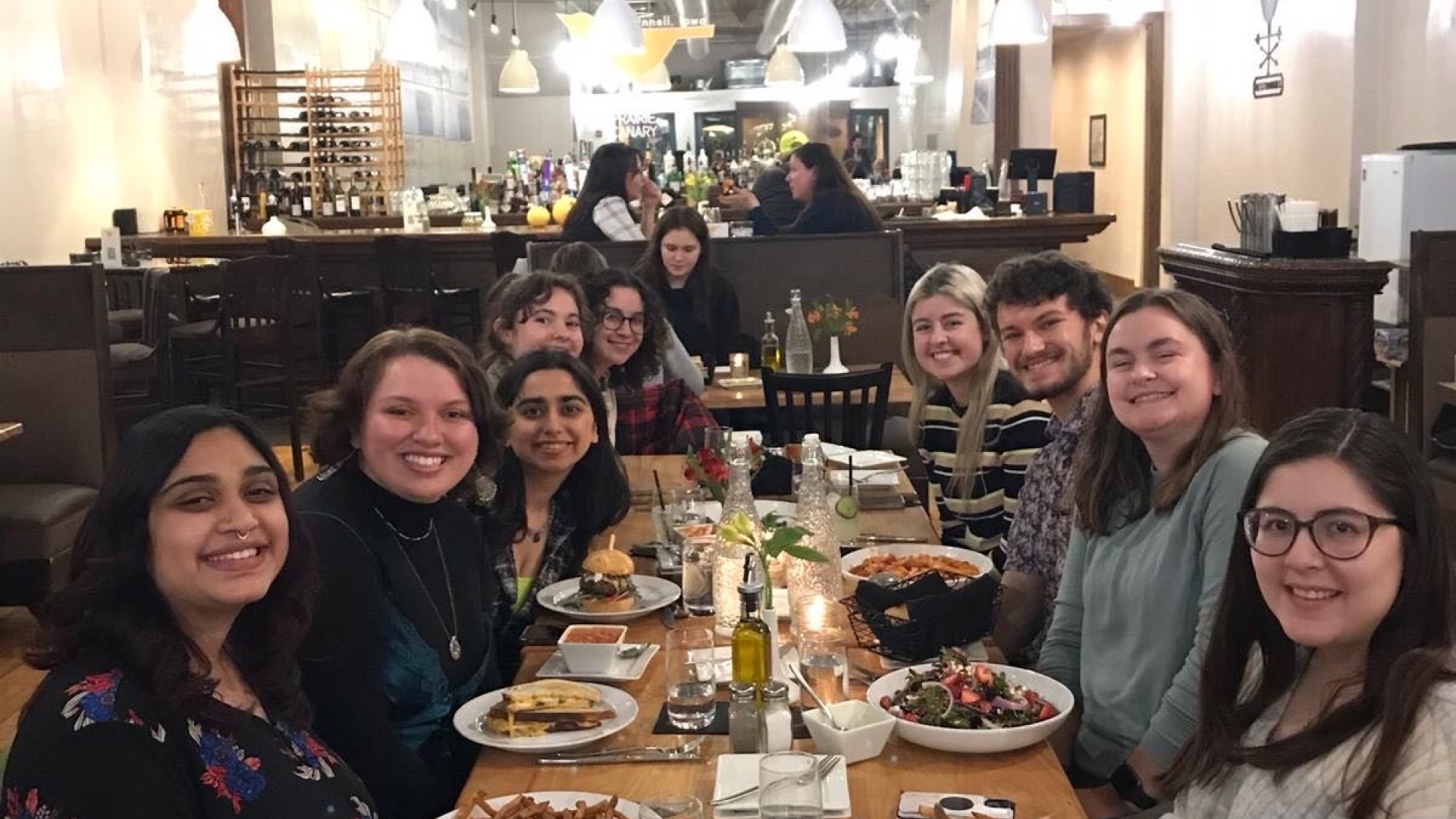 My eight friends and I all gather in front of a long wooden rectangular table in a local restaurant. We all have foods in front of us and are smiling at the camera.