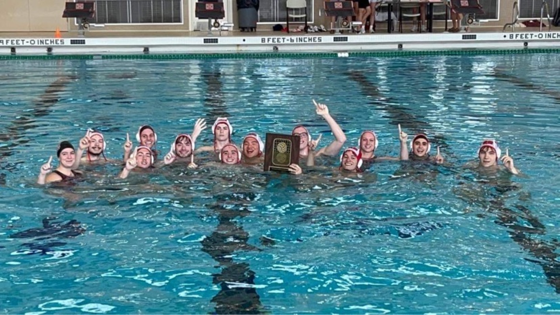 A water polo team is inside a clear swimming pool, one of the members proudly holds up in the water an award plaque in their hands.