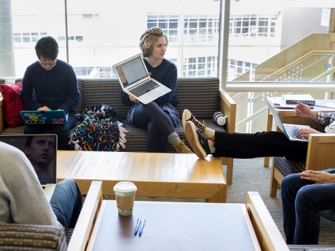 A group of students sit and type while one shows her laptop screen to another.