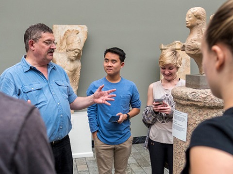 Students stand in a museum with ancient artifacts listening to a man 
