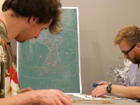 Two students work at a table with a green slate with designs in colored chalk in the background