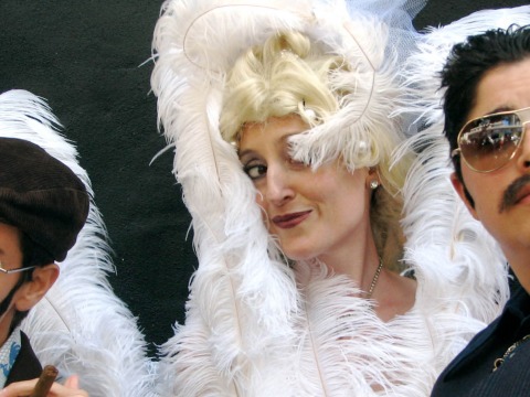 Three people: two blue-suited with mustaches and sunglasses, one with golden curls and large white feathers