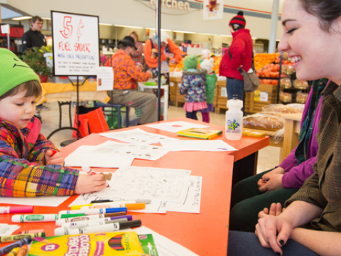 college students sit at a table in a grocery store watchinga small child coloring with markers